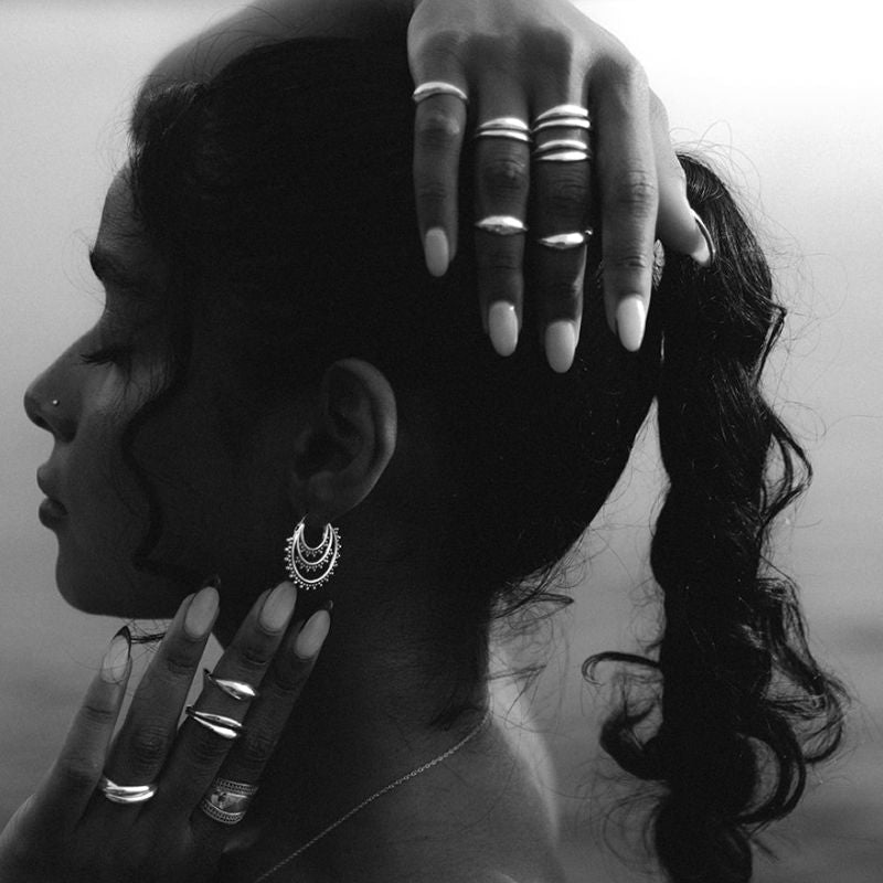 Black-and-white close-up profile of a woman with a ponytail wearing layered silver hoop earrings and multiple stacked rings, hand resting on her head.