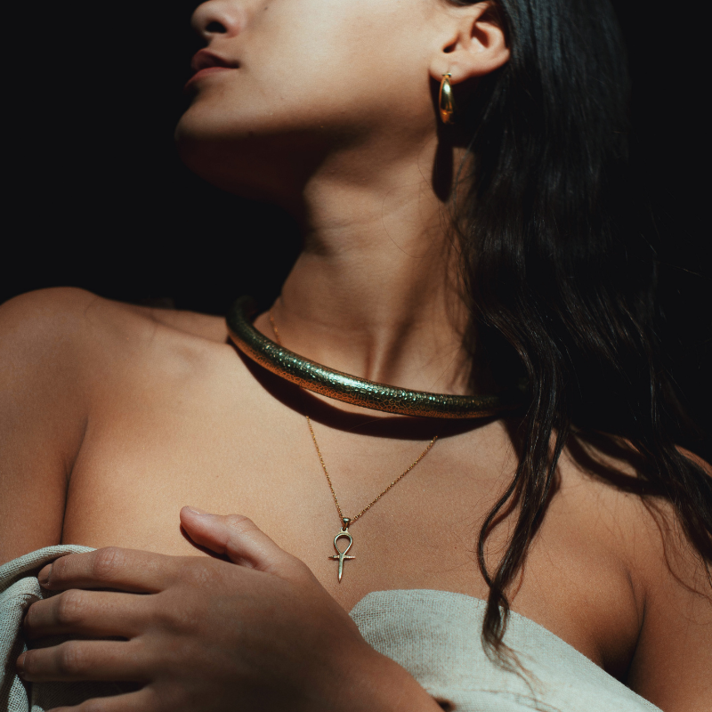 Woman wearing gold hoop earrings and layered necklaces with a symbolic pendant, styled against a dark background, elegance, and strength.