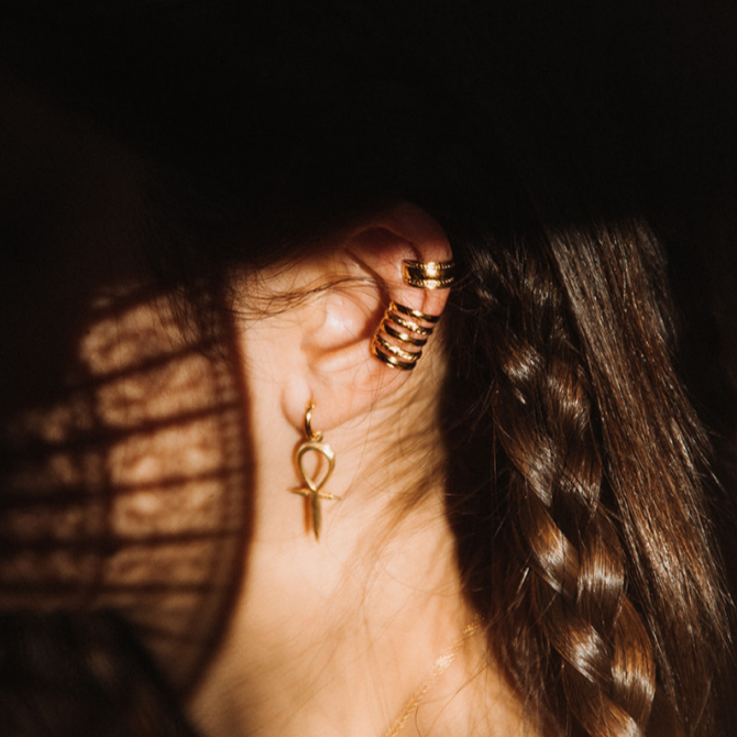 Stacked gold ear cuffs and a small gold ankh-shape charm earring photographed against dark braided hair in dramatic natural light.