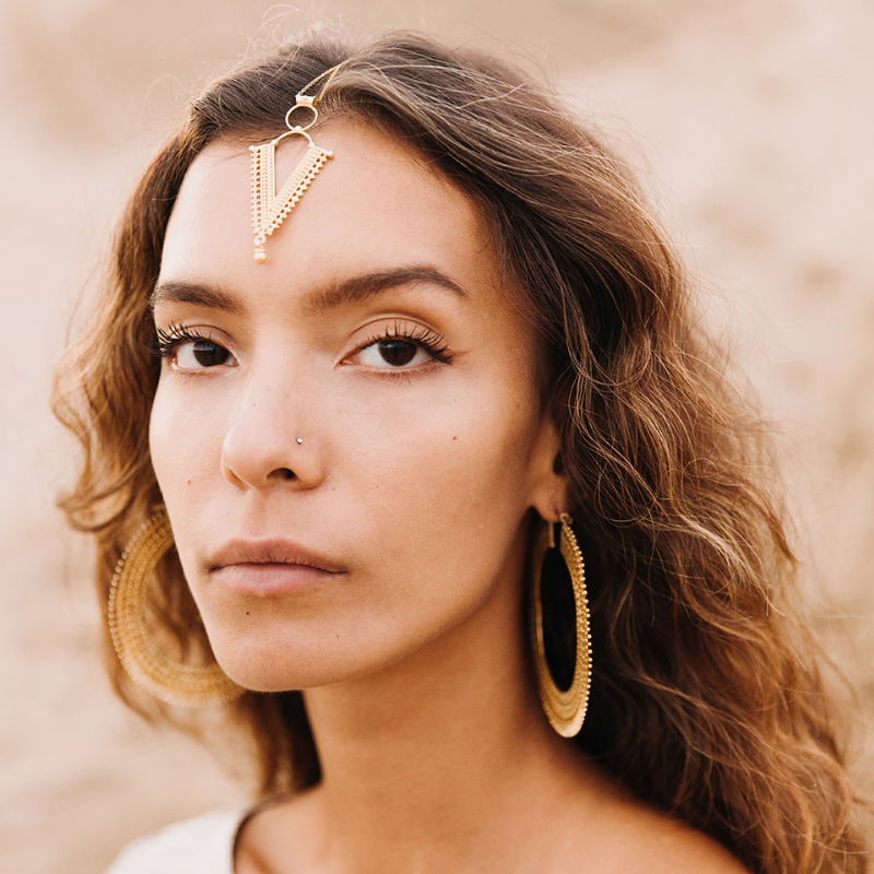 Model wearing a gold V shaped forehead chain with hoop earrings, styled against a warm neutral desert backdrop.