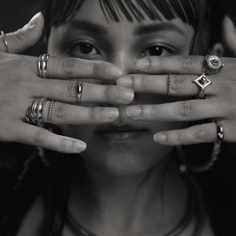 Both hands stacked with multiple 925 sterling silver rings of varied forms held over a model's face in a black and white portrait.