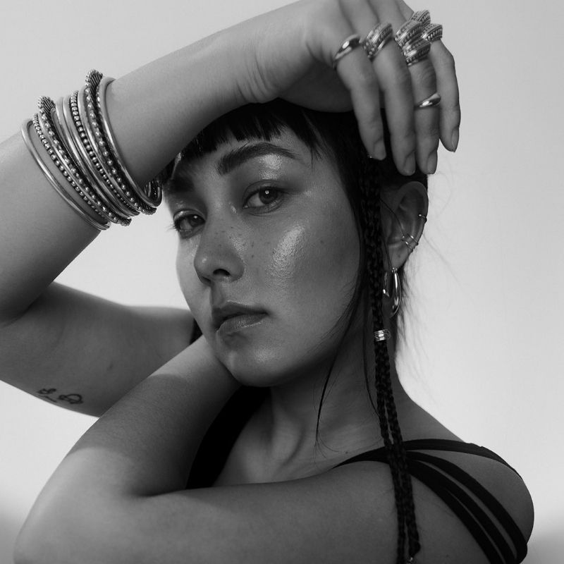 Woman with silver bangles and rings poses with arms crossed over her face, showing multiple ear piercings and a braided hair strand. - NOIR KĀLA