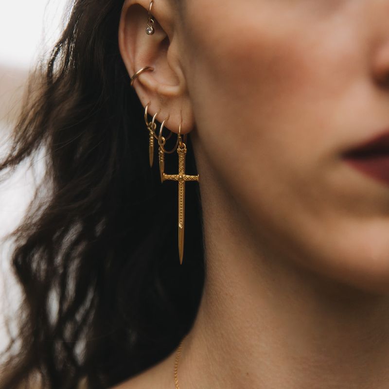 Woman wearing a layered ear stack with gold huggie earrings and engraved sword drop charms by Noir KĀLA.