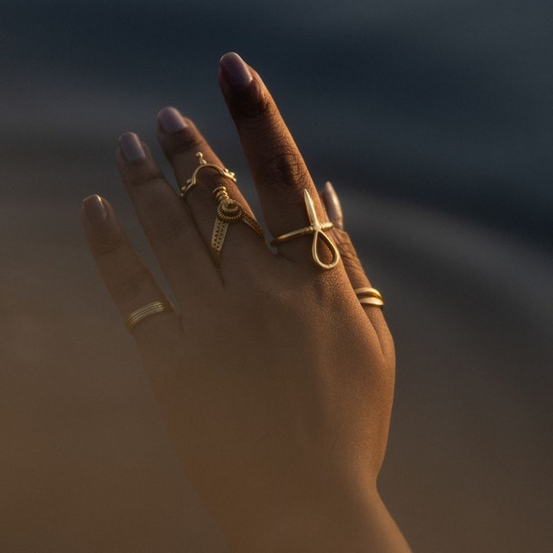 Hand wearing stacked brass rings including a sculptural knot ring, textured coil band, and filigree design in warm golden coastal light.