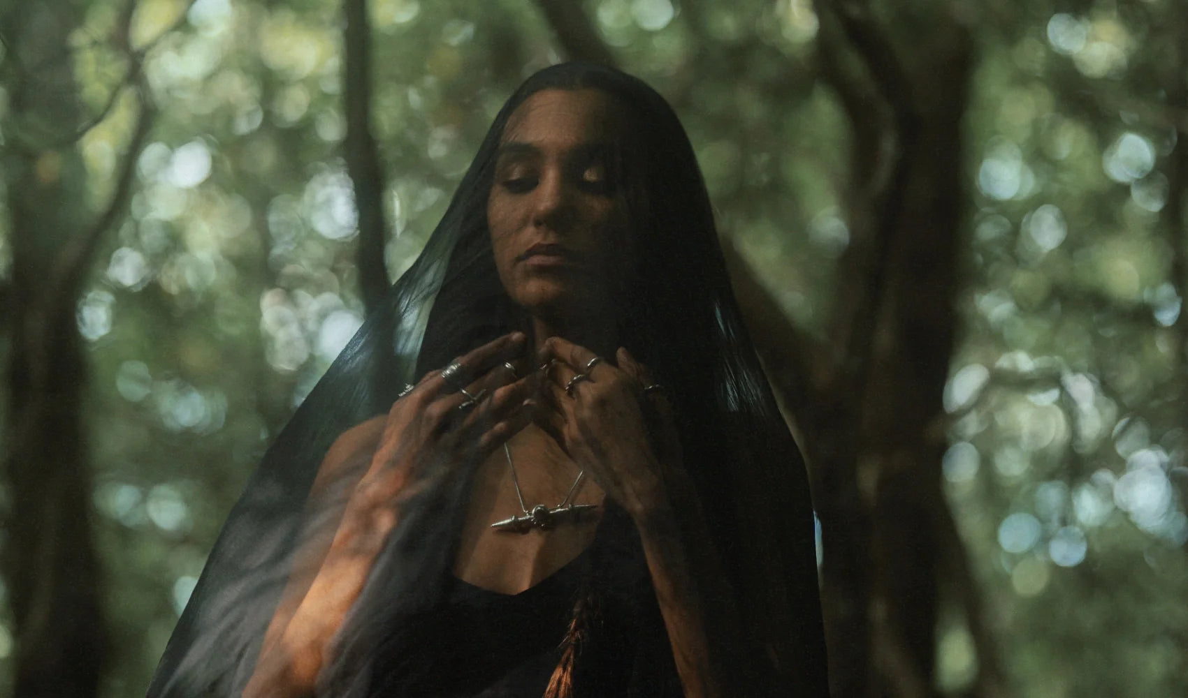 Woman wearing Noir KĀLA symbolic jewelry in a forest, layered silver rings and pendant necklace visible beneath a sheer veil, evoking ritual and mysticism.