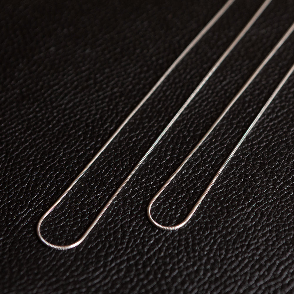 Two thin sterling silver snake chain necklaces laid in loose loops on a textured black leather surface, shown up close in detail. 