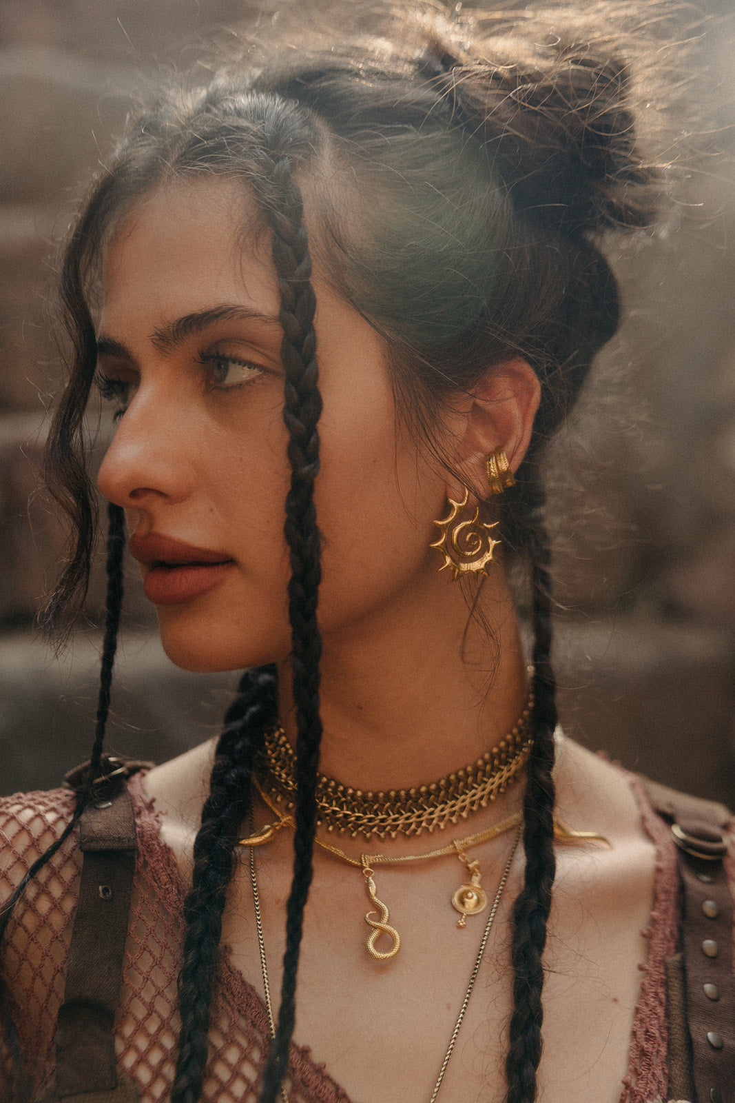 Gold sun-shaped earring styled with layered chain necklaces and braids