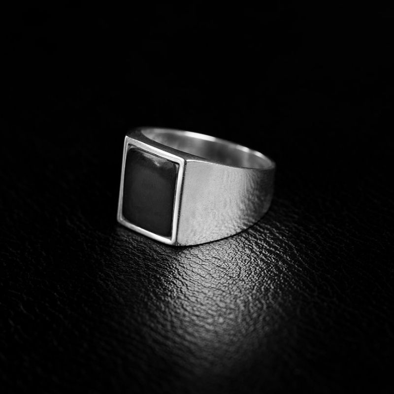 Black-and-white close-up of a square-faced sterling silver signet ring with a dark inset, resting on a textured black surface.
